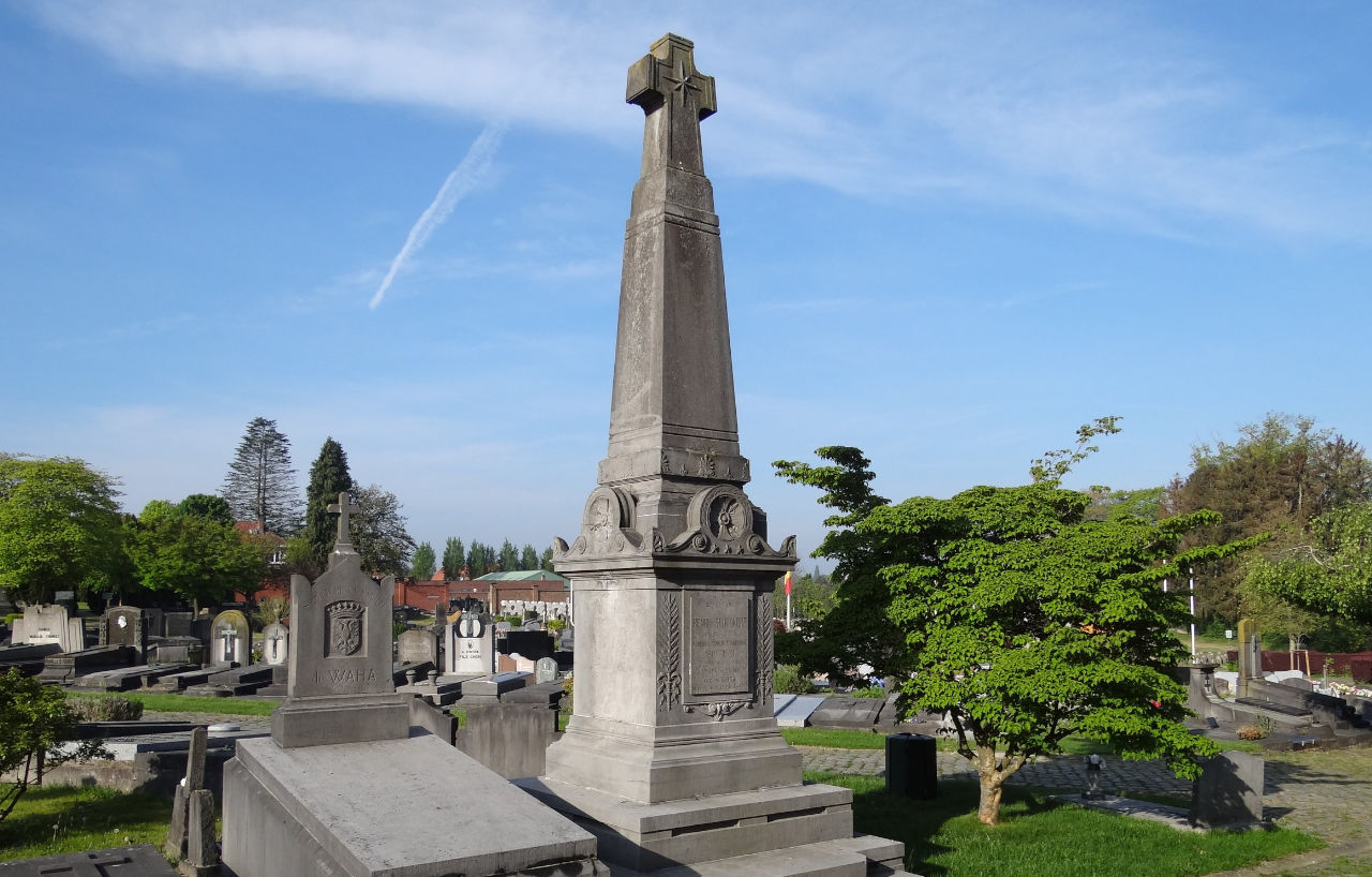 Croix de pierre à l'entrée du cimetière d'Auderghem, avec les tombes alignées derrière.