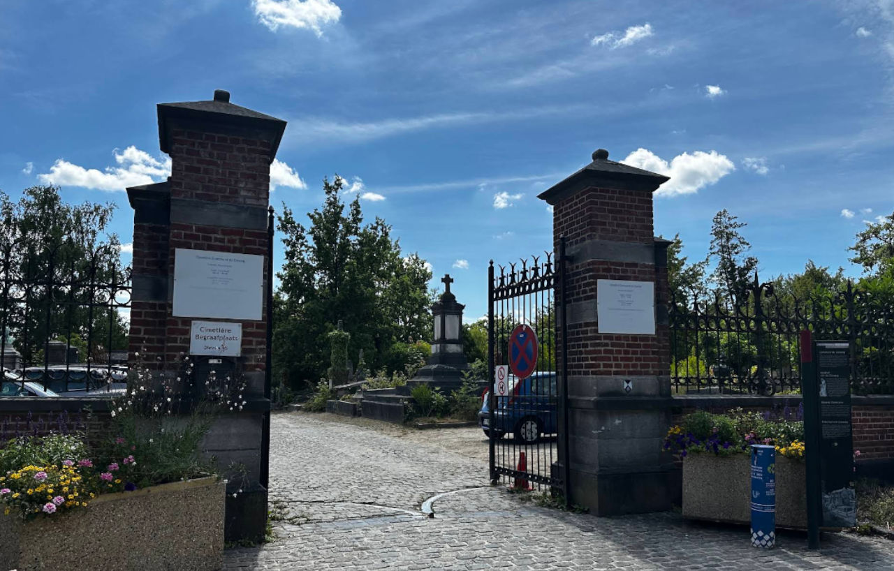 Entrée du cimetière du Dieweg à Uccle, entre deux piliers de brique, la grille ouverte sur l'allée pavée.