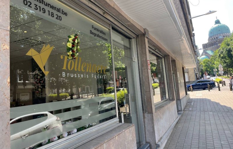 Vitrine des Funérailles Tollenaere à Anderlecht, logo à la colombe dorée et couronne de fleurs exposée.