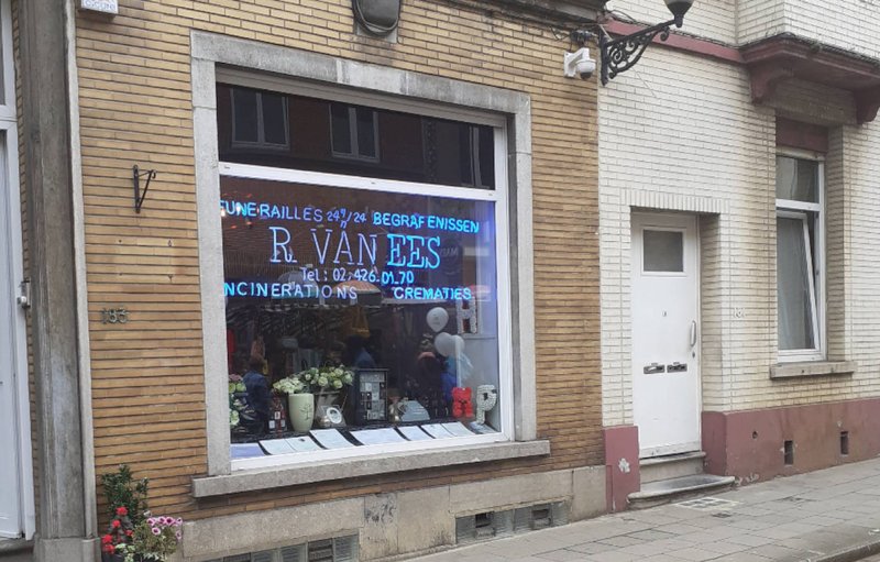 Façade de brique des Funérailles Van Ees à Jette, vitrine à lettres bleues et plantes sur le trottoir.