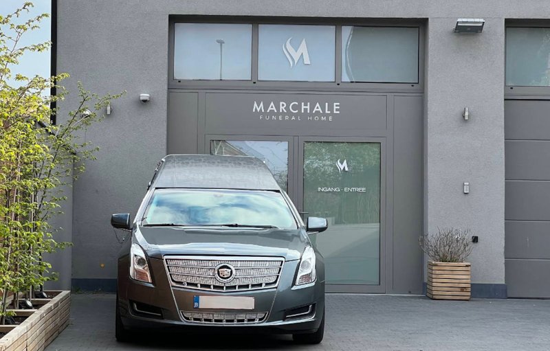Entrée du Marchale Funeral Home à Auderghem, façade grise et corbillard stationné devant la porte.